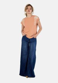 Pfirsichfarbenes Oberteil mit floralem Stickereidetail an den Schultern, kombiniert mit weiten, blauen Jeans. Glatte Textur und lässige Passform.