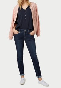 Veste bomber rose pâle texturée portée sur un chemisier bleu marine à boutons, avec un jean slim bleu foncé retroussé aux chevilles, complétée par des baskets blanches.