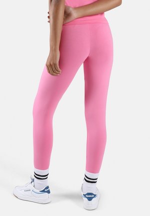 Persoon staand, gekleed in felroze leggings, witte Reebok-sneakers met blauwe accenten en witte sokken met zwarte strepen, tegen een effen achtergrond.