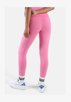 Persona de pie con mallas rosas brillantes, zapatillas blancas Reebok con detalles azules y calcetines blancos con rayas negras, sobre un fondo liso.