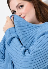 Blaues Strickpullover mit gerippter Textur, locker sitzend mit weiten Ärmeln. Der Stoff wirkt weich und gemütlich, gerafft am Ausschnitt.