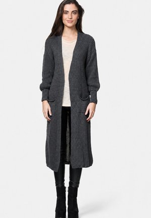 Vrouw staand, gekleed in een lange donkergrijze gebreide cardigan met zakken, een lichtbeige blouse, zwarte broek en zwarte enkellaarzen.