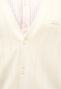 Κρέμα καζάκ με κάθετη ραβδωτή υφή, σχέδιο σε V και τρία κουμπιά. Επικαλυμμένο με ένα ανοιχτό ροζ και λευκό ριγέ πουκάμισο.