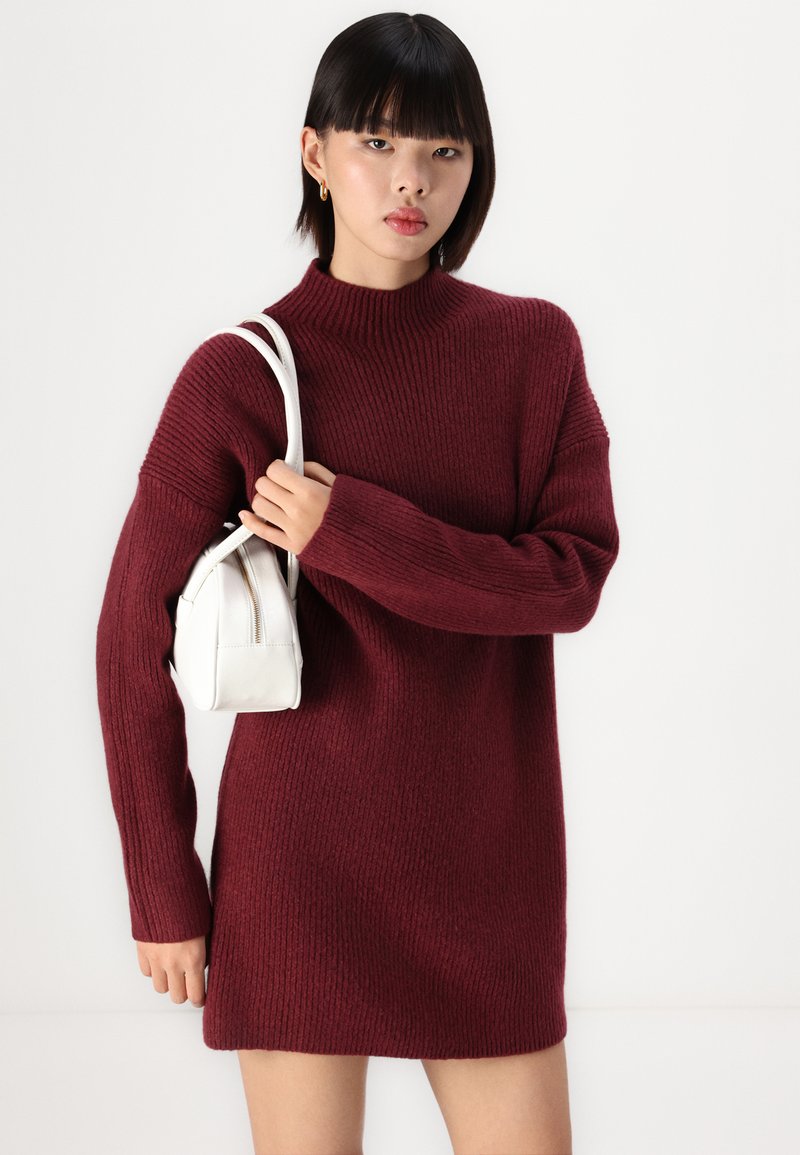 Bordeaux gestricktes Kleid mit hohem Kragen, geripptem Muster und langen Ärmeln. Das Model hält eine kleine weiße Handtasche.