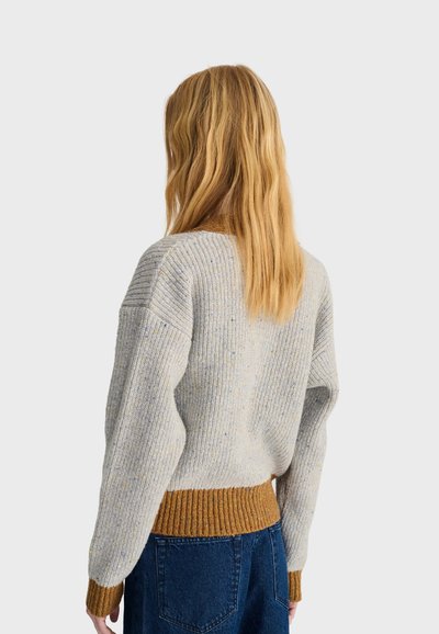 Femme aux longs cheveux blonds porte un pull en maille gris clair avec des poignets et un ourlet côtelés marron, associé à un jean bleu foncé, tournée de dos.
