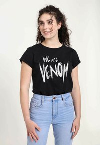 Černé tričko s krátkým rukávem s výrazným bílým grafickým textem "JSME VENOM." V kombinaci se světle modrými džíny s vysokým pasem.