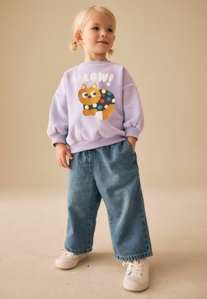 Lila Sweatshirt mit einem Cartoon-Katzenmotiv und dem Schriftzug "MEOW!" kombiniert mit locker sitzenden blauen Jeans und weißen Turnschuhen.