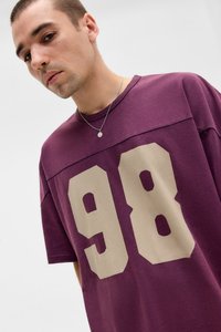 Jonge man met kort haar, gekleed in een paarse shirt met een grote beige "98" op de voorkant, een kleine hanger om zijn nek en een kleine ringoorbel, kijkt naar de camera.
