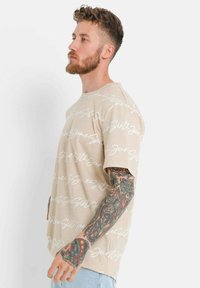 Beige t-skjorte med kort erme og hvit skrift. Løs passform, rund hals og strukturert stoff. Merker fra tatoveringer er synlige på armen.