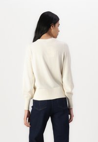 Pull en tricot de couleur crème avec des poignets et un ourlet côtelés, des manches bouffantes et une coupe décontractée. Porté avec un jean bleu foncé.