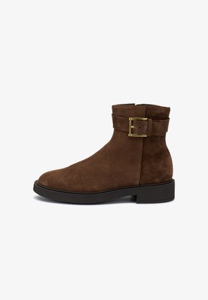 Bottines en suède marron avec un bout arrondi, une semelle noire plate et un accent en boucle dorée. Présente une texture lisse et une hauteur jusqu'au mollet.
