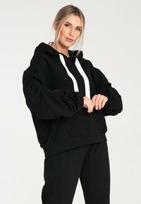 Czarna bluza z kapturem wykonana z miękkiego materiału, z białymi sznurkami, z marszczonymi rękawami i luźnym krojem. W połączeniu z pasującymi czarnymi spodniami.