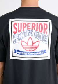T-shirt en coton noir avec un grand motif blanc au dos, accompagné du texte rouge et bleu "SUPERIOR SPORTING GOODS" et du logo Adidas.