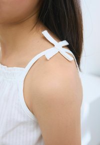 White sleeveless top with a ruffled neckline, featuring narrow straps tied in bows on the shoulders, showcasing a smooth fabric texture.