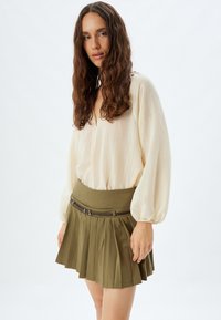 Blouse crème à manches longues avec une texture douce, associée à une jupe plissée vert olive dotée d'une large ceinture et d'accents en cuir marron.