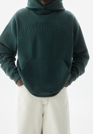 Person trägt dunkelgrünen Hoodie mit eingeprägtem Schriftzug "NEW YORK CITY" auf der Vorderseite, kombiniert mit hellbeigen Hosen, Hände in den Taschen.