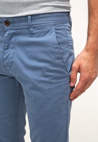 Ljust blå bomullschinos med slim fit, med sidofickor och en synlig brun knapp i midjan. Slät текстur och subtila sömmar.