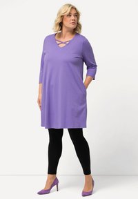 Vestito tunica viola, lungo fino al ginocchio, con maniche a tre quarti e scollatura incrociata, abbinato a leggings neri e tacchi alti viola.