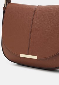 Brun tekstureret crossbody taske med en rund flap, guld brandingplade og kontrastsyninger. Den har en glat, holdbar læderfinish.