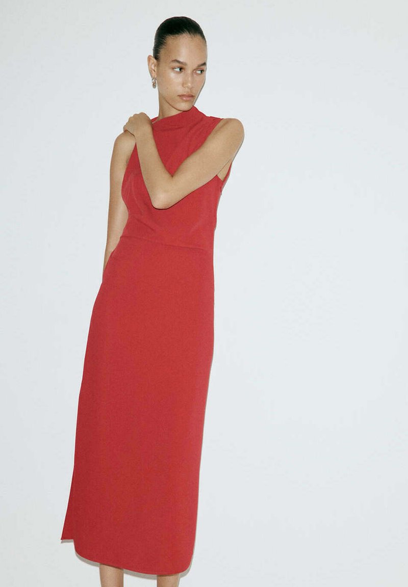 Femme aux cheveux foncés portant une robe midi rouge sans manches, debout devant un fond blanc uni, une main posée sur son épaule.