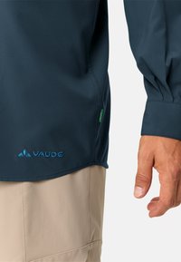 Giacca esterna blu scuro con finitura testurizzata, con logo Vaude azzurro chiaro, fessure laterali per ventilazione e polsini elasticizzati.