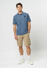 Vīriešu zils polo krekls ar īsām piedurknēm, ar logo uz krūtīm, apvienots ar bēšiem cargo šortiem un baltām sporta zeķēm un apaviem.