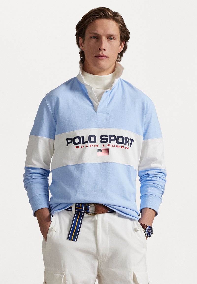 Gaiši zils garām piedurknēm krekls ar baltu horizontālu svītru, kurā redzams teksts "POLO SPORT" tumši zilā un sarkanā krāsā, uzvilkts virs balta turtlēka.