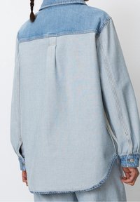 Vaaleansininen farkkupaita, jossa on tummansininen yoke, pitkät hihat, sivusaumat ja kaareva helma. Ominaisuuksiin kuuluu takakappaleen laskos ja napilliset hihansuut.