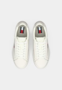 O pereche de adidași albi Tommy Jeans cu șireturi, cu logo-ul vizibil pe tălpi și laturi, expuși din vedere de sus pe fundal alb.