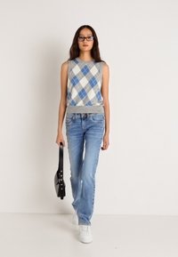 Γκρι αμάνικο πουλόβερ με μπλε και λευκό μοτίβο αργυλής, συνδυασμένο με ανοιχτό γαλάζιο τζιν και λευκά αθλητικά παπούτσια. Κρατάει μια μαύρη τσάντα χειρός.