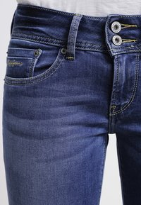 Mörkblå denimjeans med slim fit, prydda med två metallknappar, kontrasterande gul sömnad och klassisk femficksdesign.
