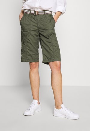 Person iført knælange olivengrønne cargoshorts, et stribet bælte, hvide sneakers og en hvid skjorte med hænderne i lommerne.