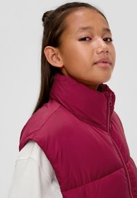 Lila ärmellose Steppjacke mit hohem Kragen und Reißverschluss, getragen über einem weißen Langarmshirt. Glatte Textur und leichtes Design.