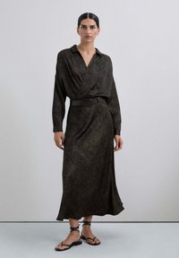 Vestido largo de color marrón oscuro en tejido texturizado con cuello, mangas largas y cinturón ancho. Hebilla de cinturón circular y dobladillo fluido. Sandalias planas.