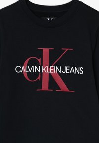 Sudadera negra con texto en negrita: "CALVIN KLEIN JEANS" con una "K" roja prominente y una "c" más pequeña en rojo.