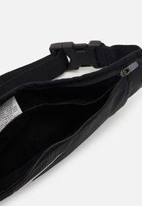 Bolsa de cinturón de nailon negro con correa ajustable, compartimento principal con cremallera, etiqueta interior y detalle de logo brillante. Textura suave y diseño ligero.