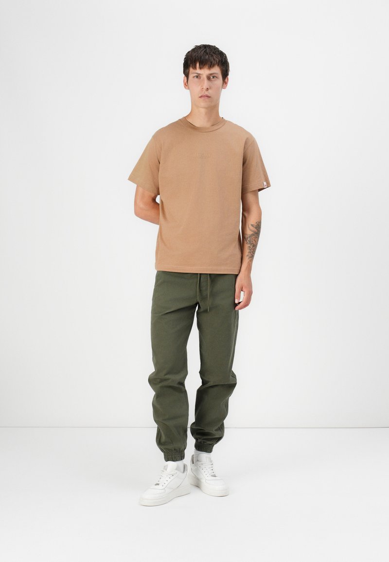 T-shirt in cotone marrone, maniche corte, collo rotondo; abbinato a pantaloni verde oliva, con polsini elastici e sneakers bianche con suola spessa.