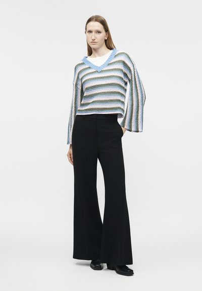 Femme debout, la main droite dans la poche, portant un pull à col en V rayé bleu, blanc et vert, un pantalon large noir et des chaussures noires.