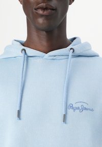 Gaiši zils kapuci krekls ar auklu un sudraba aksesuāriem. Uz krūtīm ir izšūts "Pepe Jeans" logo tumšākā zilā krāsā.