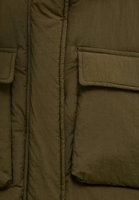Olivegrüne Pufferjacke mit strukturiertem Finish, ausgestattet mit großen, umklappbaren Fronttaschen und sichtbaren Nähten entlang der Nähte.