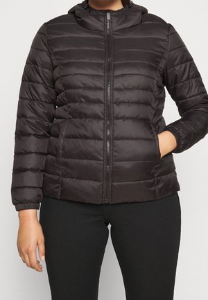 Person trägt eine schwarze gesteppte Daunenjacke mit Frontreißverschluss und Kapuze, eine Hand in der Tasche, kombiniert mit schwarzen Hosen.