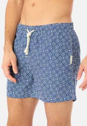 Buste masculin portant un short de bain bleu avec un motif géométrique blanc et un cordon blanc, sur un fond clair uni.