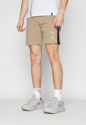 Beige sportshorts lavet af letvægtsstof, med sorte accenter på siden og grafiske designs foran. Båret med grå sneakers og hvide sokker.