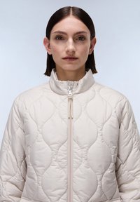 Gegolfde, lichtbeige jas met een hoge kraag. Voorzien van een ritssluiting aan de voorkant en een gladde, glanzende textuur met een golvenpatroon.
