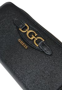 Cartera negra texturizada con un logo en tono dorado y un acento de cadena. Cierre con cremallera con una etiqueta de cuero; forma rectangular compacta.