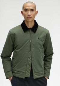 Giacca verde con zip, colletto in velluto a coste nero, tasche frontali e logo sul lato sinistro del petto. Tessuto liscio, design moderno.