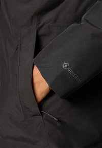 Crna Gore-Tex jakna s glatkom teksturom, šavovima na sebi i vidljivim logotipom na rukavu. Džep vidljiv na struku.