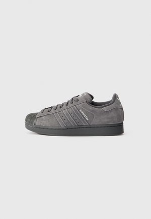 Grauer Adidas Superstar Sneaker mit schwarzer Gummikappe, Wildlederobermaterial, drei Streifen und gebrandeten Details an der Seite und Ferse.