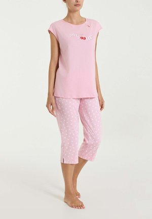 Donna con maglietta rosa a maniche corte con disegno di ciliegie e pantaloni pigiama rosa corti abbinati con motivo floreale bianco. A piedi nudi.