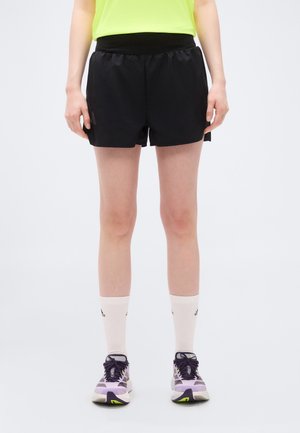 Persoon draagt zwarte sportshorts, witte sokken tot halverwege de kuit met logo's, en paarse hardloopschoenen, staand op een effen witte achtergrond.
